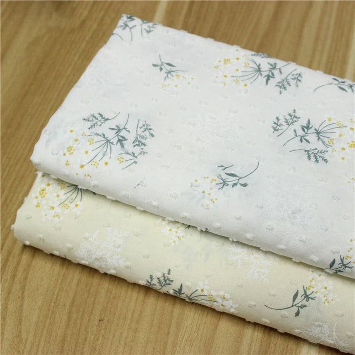 Deux pièces pliées du Tissu plumetis fleuri au mètre de Longan Craft France, l'une blanche et l'autre beige clair avec des imprimés floraux verts et jaunes, présentées sur une surface en bois.