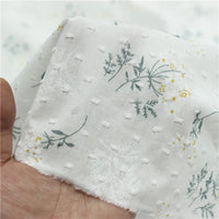 Une main tient le Tissu plumetis fleuri au mètre de Longan Craft France, un tissu de coton blanc et jaune à la texture pointillée, avec des feuilles vertes, des tiges et de petites fleurs jaunes.