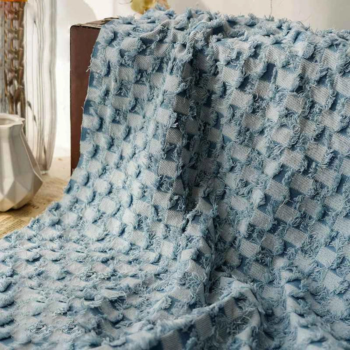 Un morceau de Tissu denim à franges au mètre Longan Craft France, présentant un motif à carreaux et des bords effilochés, est drapé sur une surface à côté d'un vase blanc et d'un récipient en verre pour une touche décorative polyvalente.