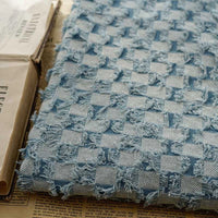 Gros plan sur le Tissu denim à franges au mètre de Longan Craft France, un tissu jean effiloché décoratif bleu et blanc, posé sur un livre ouvert et un journal.