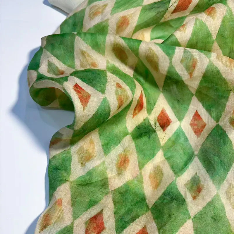Un morceau de Tissu en ramie à imprimé géométrique au mètre de Longan Craft France, présentant un motif en losange dans les tons vert, crème, orange et jaune, est drapé sur une surface. Ce tissu léger et semi-transparent est idéal pour les robes d'été.