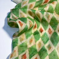 Un morceau de Tissu en ramie à imprimé géométrique au mètre de Longan Craft France, présentant un motif en losange dans les tons vert, crème, orange et jaune, est drapé sur une surface. Ce tissu léger et semi-transparent est idéal pour les robes d'été.