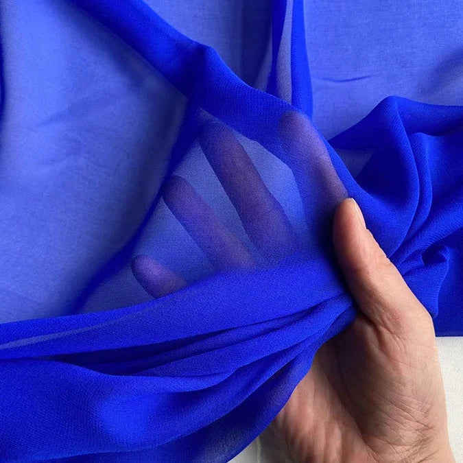 Une main soulève la Georgette de soie au mètre de Longan Craft France, un crêpe de soie 100% mûrier (8 mommes) bleu vif et transparent, révélant sa texture légère et transparente et son drapé fluide alors que les doigts sont visibles à travers le tissu.
