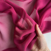 Une main brandit le Georgette de soie au mètre de Longan Craft France - un crêpe de soie 100 % mûrier, 8 mamans - qui montre sa couleur rose vif diaphane et son drapé fluide lorsque la lumière passe à travers le tissu léger.