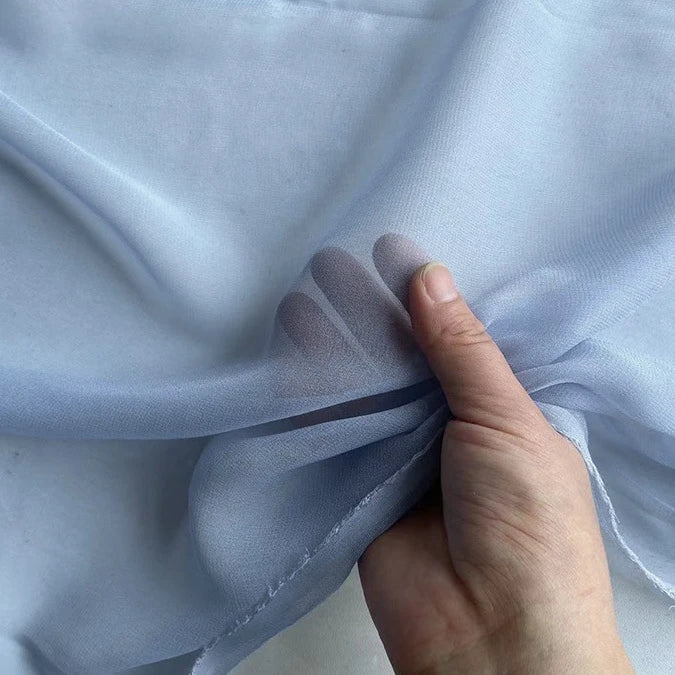 Une main présente une Georgette de soie au mètre bleu clair semi-transparente de Longan Craft France, un crêpe souple 100 % soie de mûrier (8 mamans) au drapé délicat et aux doigts visibles à travers le tissu.