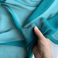 Une main fronce doucement la Georgette de soie au mètre de Longan Craft France - ce tissu 100% soie de mûrier, 8 mommes, est fluide et translucide, révélant le contour subtil des doigts en dessous.