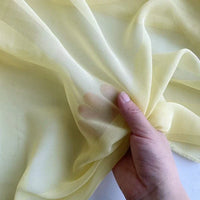 Une main tient la Georgette de soie au mètre de Longan Craft France, un crêpe diaphane jaune clair 100 % soie de mûrier (8 mamans). Le tissu souple est légèrement froncé, montrant son drapé fluide et sa qualité transparente.