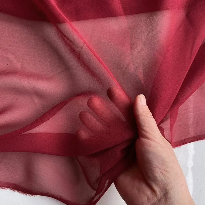 Une main tient un morceau de Georgette de soie au mètre de Longan Craft France, 100% mûrier crêpe de soie (8 mommes), tirant légèrement le tissu de soie léger et transparent pour révéler son drapé fluide et sa transparence sur un fond blanc.
