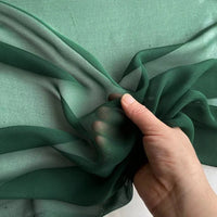 Une main tient la Georgette de soie au mètre de Longan Craft France - crêpe fluide 100 % soie de mûrier, 8 mamans - en vert foncé transparent sur un fond plus clair, montrant le drapé délicat et la semi-transparence de l'étoffe.