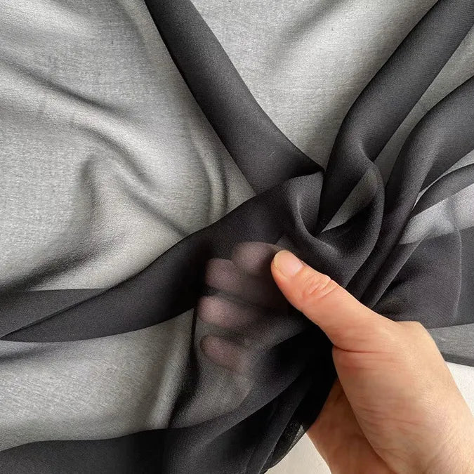 Une main tient et froisse la Georgette de soie au mètre de Longan Craft France-un tissu en soie 100% mûrier, 8 mommes- sur un fond gris, mettant en valeur sa texture souple et fluide et sa belle transparence.