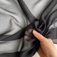 Une main tient et froisse la Georgette de soie au mètre de Longan Craft France-un tissu en soie 100% mûrier, 8 mommes- sur un fond gris, mettant en valeur sa texture souple et fluide et sa belle transparence.