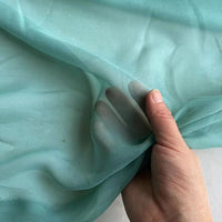 Une main brandit la Georgette de soie au mètre-crêpe de soie 100% mûrier, 8 mommes, de Longan Craft France. Le tissu léger et transparent est délicat et fluide, laissant apparaître les doigts à travers son drapé transparent.