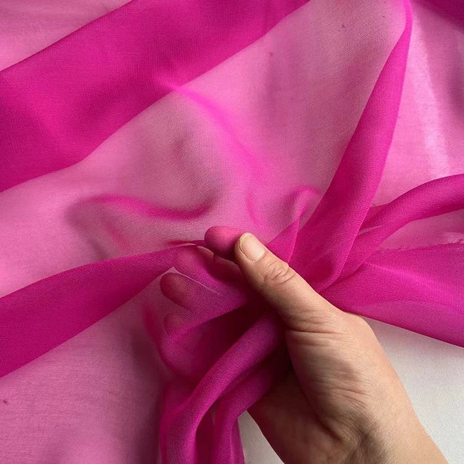 Une main tient la Georgette de soie au mètre de Longan Craft France, un tissu diaphane et rose vif 100 % soie de mûrier (8 mommes), mettant en valeur son drapé doux et fluide et sa texture transparente sur un fond assorti.