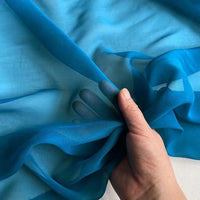 Une main tient un morceau de Georgette de soie au mètre de Longan Craft France, un tissu transparent et léger 100 % soie de mûrier (8 mamans), révélant son drapé fluide et sa texture douce grâce à sa qualité semi-transparente.