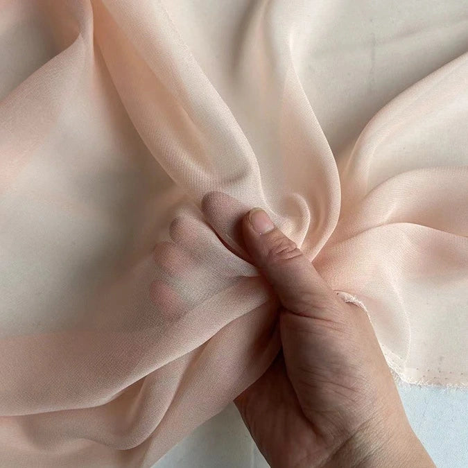 Une main saisit la Georgette de soie au mètre de Longan Craft France - 100 % soie de mûrier, 8 mamans - un tissu crêpe de soie transparent, léger et fluide, doucement froncé pour mettre en valeur son drapé délicat sur un fond neutre.