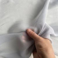 Une main soulève un morceau de Georgette de soie au mètre de Longan Craft France - un tissu en soie 100% mûrier, 8 mommes, souple et fluide. Le crêpe diaphane gris clair apparaît semi-transparent et délicat sur un fond uni.