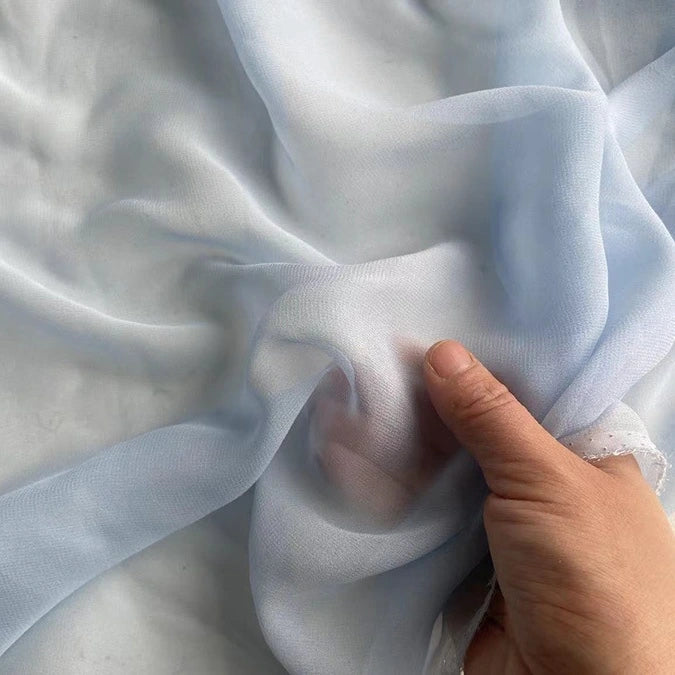 Une main fronce la Georgette de soie au mètre bleu clair de Longan Craft France, un crêpe 100 % soie de mûrier de 8 mm d'épaisseur, doux, léger et fluide pour un drapé élégant.