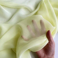 Une main se presse contre une Georgette de soie au mètre jaune pâle de Longan Craft France, les doigts étant visibles à travers le tissu 100 % soie de mûrier. Ce crêpe de soie 8 momme, doux et fluide, se drape délicatement sur la majeure partie de la main.