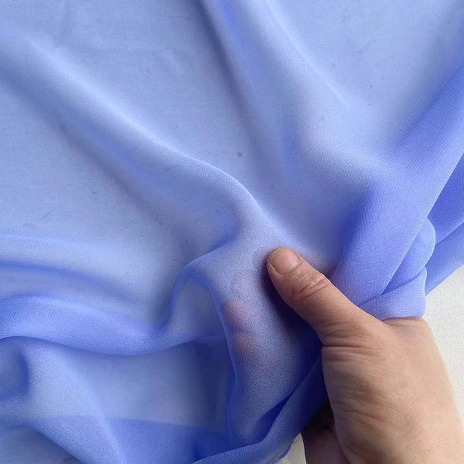 Une main tient et rassemble un morceau de Georgette de soie au mètre de Longan Craft France, un crêpe de soie 100 % mûrier (8 mommes) bleu clair, montrant sa texture transparente, douce et fluide, idéale pour le drapé.