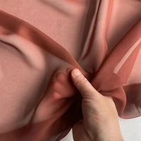 Une main rassemble le Georgette de soie au mètre de Longan Craft France, un crêpe de soie 100 % mûrier (8 mommes), transparent et brun-rouge, révélant le contour de la main à travers le drapé semi-transparent, doux et fluide, sur un fond clair.