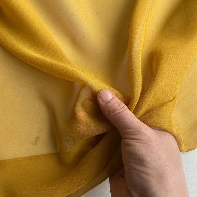 Une main rassemble un morceau de Georgette de soie au mètre de Longan Craft France, un tissu transparent et léger 100 % soie de mûrier (8 mommes), mettant en valeur son drapé transparent, doux et fluide.
