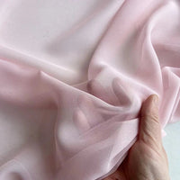 Une main tient et froisse délicatement la Georgette de soie au mètre de Longan Craft France, un tissu doux et fluide 100 % soie de mûrier (8 mommes), révélant son drapé délicat et sa texture diaphane sur un fond uni.