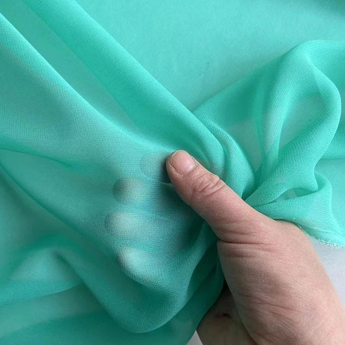 Une main pince la "Georgette de soie au mètre" translucide et légèrement turquoise de Longan Craft France, un crêpe 100 % soie de mûrier (8 mommes) qui met en valeur son drapé diaphane et fluide ; l'arrière-plan présente le même tissu délicat.
