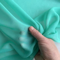 Une main pince la "Georgette de soie au mètre" translucide et légèrement turquoise de Longan Craft France, un crêpe 100 % soie de mûrier (8 mommes) qui met en valeur son drapé diaphane et fluide ; l'arrière-plan présente le même tissu délicat.