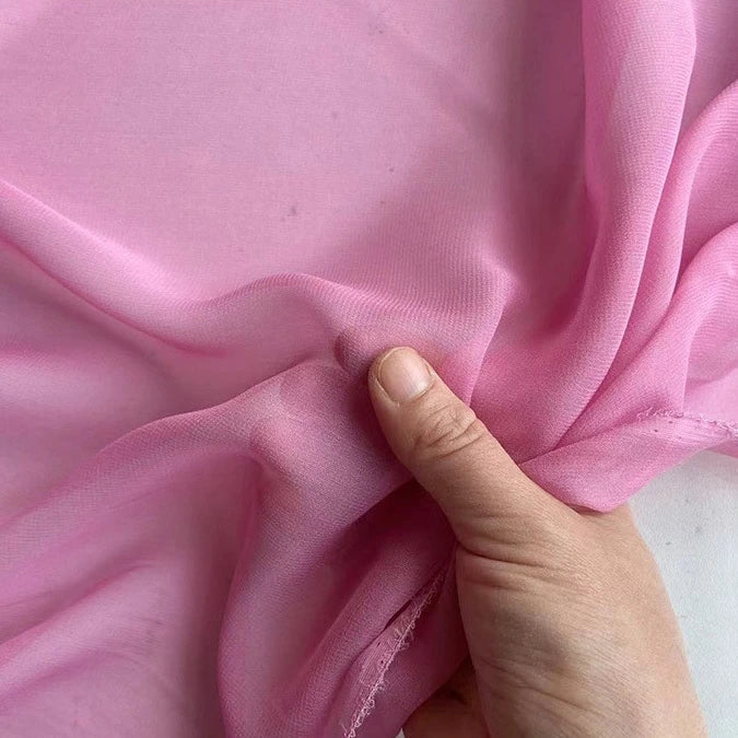 Une main tient la Georgette de soie au mètre de Longan Craft France, 100% crêpe de soie de mûrier (8 mommes), montrant la translucidité rose pâle de ce tissu doux, souple et fluide, idéal pour le drapé.