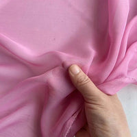 Une main tient la Georgette de soie au mètre de Longan Craft France, 100% crêpe de soie de mûrier (8 mommes), montrant la translucidité rose pâle de ce tissu doux, souple et fluide, idéal pour le drapé.