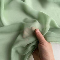 Une main tient et froisse délicatement la Georgette de soie au mètre de Longan Craft France, un tissu vert clair, transparent, 100 % soie mûrier (8 mommes), doux, délicat et parfait pour des drapés fluides.