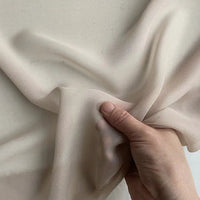 Une main tient délicatement la Georgette de soie au mètre de Longan Craft France, déroulant le crêpe de soie 100 % mûrier (8 mommes), léger et transparent, pour révéler des plis doux et des ombres subtiles sur un fond uni.