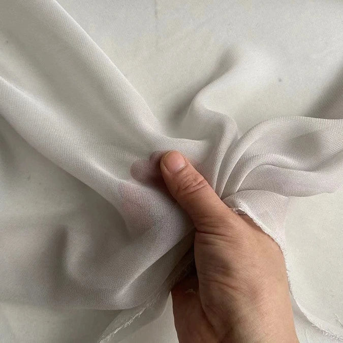 Une main soulève un morceau de Georgette de soie au mètre de Longan Craft France, un crêpe de soie 100 % mûrier de 8 mm. Le tissu est doux, semi-transparent et se drape avec fluidité sur un fond blanc.