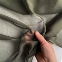 Une main tient et fronce la Georgette de soie au mètre de Longan Craft France, un tissu transparent vert olive 100 % soie de mûrier (8 mamans), mettant en valeur son drapé léger, translucide et fluide.