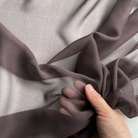 Une main tient une Georgette de soie au mètre transparente brun foncé de Longan Craft France - 100 % soie de mûrier, 8 mamans - sur un fond gris clair, mettant en valeur le drapé fluide et la texture transparente de l'étoffe.