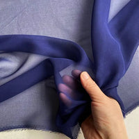 Une main froisse délicatement une Georgette de soie au mètre bleu foncé de Longan Craft France - 100 % soie de mûrier, 8 mamans - pour en révéler la transparence et la texture fluide, idéale pour les drapés.