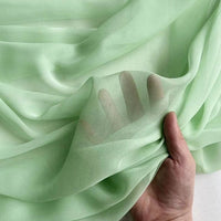 Une main fronce la Georgette de soie au mètre vert clair de Longan Craft France - ce crêpe 100% soie de mûrier (8 mommes) est translucide et fluide, dévoilant délicatement les doigts grâce à son drapé élégant.