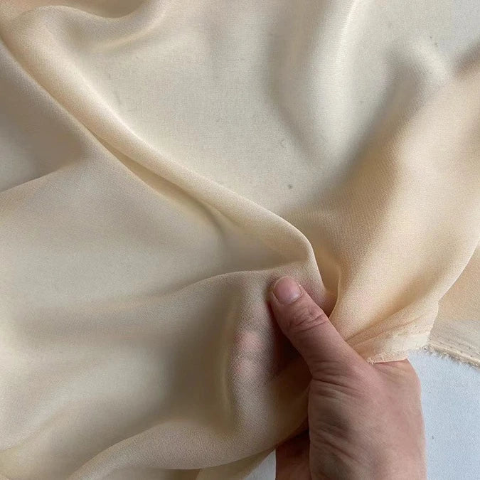Une main présente le Georgette de soie au mètre de Longan Craft France, en déroulant le crêpe de soie 100% mûrier (8 mommes), léger et semi-transparent, pour mettre en valeur sa texture souple et fluide et son beau drapé sur un fond uni.