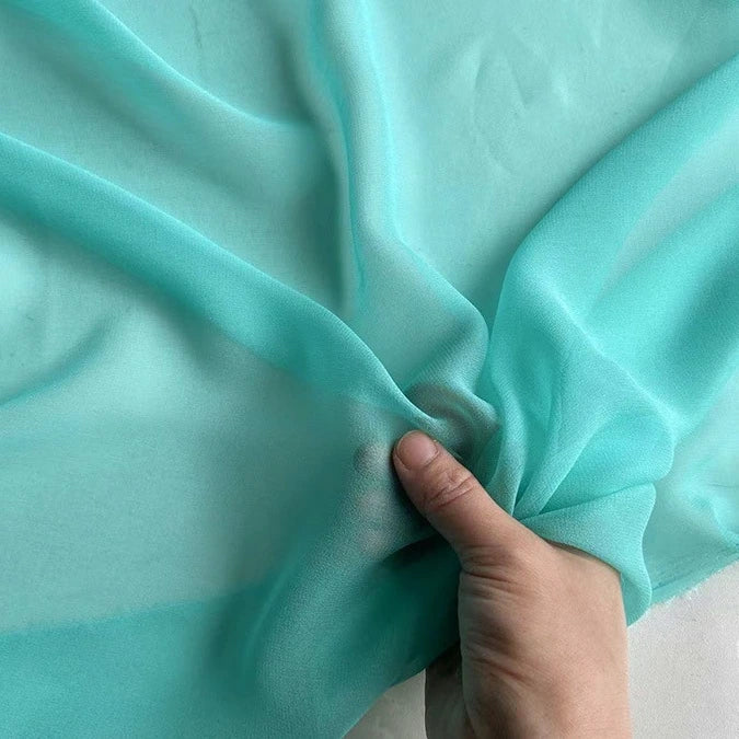 Une main tient un morceau de Georgette de soie au mètre de Longan Craft France, révélant la légèreté et la translucidité du tissu 100 % soie de mûrier, idéal pour le drapé grâce à sa texture souple et fluide.