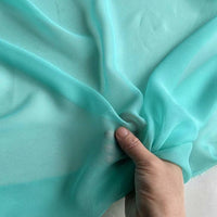 Une main tient un morceau de Georgette de soie au mètre de Longan Craft France, révélant la légèreté et la translucidité du tissu 100 % soie de mûrier, idéal pour le drapé grâce à sa texture souple et fluide.