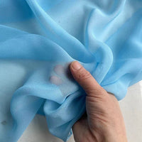 Une main rassemble un morceau de Georgette de soie au mètre de Longan Craft France, un crêpe 100% soie de mûrier (8 mommes) bleu clair, transparent et délicat, montrant son drapé transparent et fluide. Les doigts sont visibles à travers la finesse de la matière.