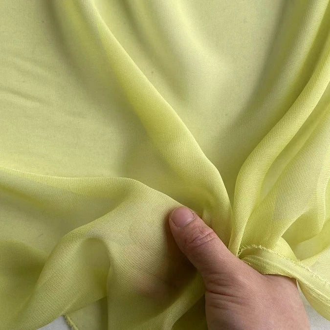 Une main présente la Georgette de soie au mètre semi-transparente et jaune clair de Longan Craft France, un tissu 100 % soie de mûrier (8 mommes) connu pour son drapé souple, sa texture délicate et sa brillance subtile.