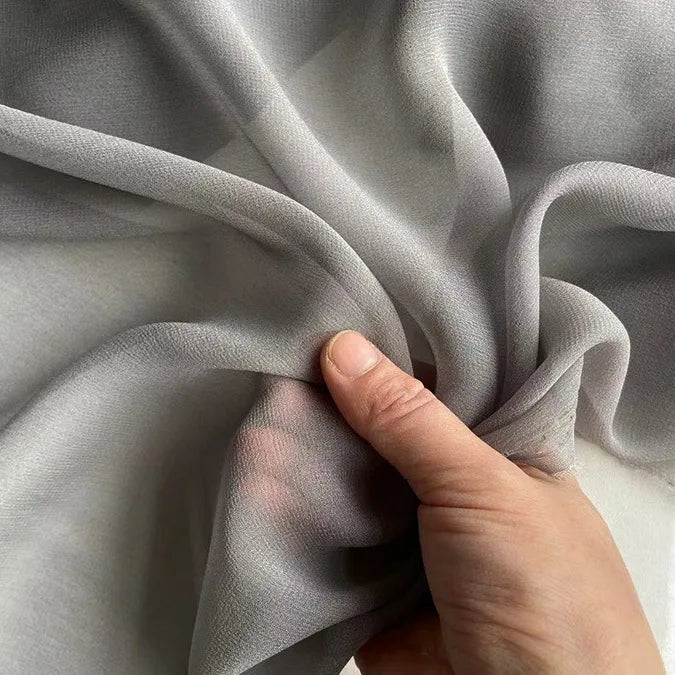 Une main rassemble délicatement un morceau de Georgette de soie au mètre de Longan Craft France, un crêpe de soie 100 % mûrier (8 mommes) gris clair et semi-transparent, formant des plis délicats dans ce tissu de soie doux et fluide, idéal pour les drapés.