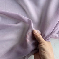 Une main pince et soulève la Georgette de soie au mètre de Longan Craft France, un tissu diaphane 100 % soie de mûrier (8 mamans), mettant en valeur sa translucidité et son drapé doux et fluide sur un fond neutre.