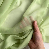 Une main tient et regroupe la Georgette de soie au mètre vert clair de Longan Craft France, un tissu diaphane 100 % soie de mûrier (8 mommes) au drapé doux et à la fluidité visible à la main grâce à sa texture translucide.
