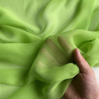 Une main tient et regroupe délicatement la Georgette de soie au mètre de Longan Craft France, une étoffe transparente 100 % soie de mûrier (8 mommes) vert clair au drapé doux, dont la translucidité laisse transparaître les doigts.