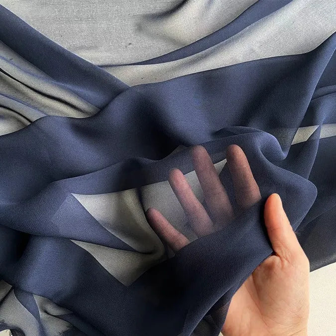 Une main présente le Georgette de soie au mètre de Longan Craft France - un drapé en crêpe de soie 100% mûrier, 8 mommes - révélant sa texture diaphane et fluide ainsi que sa translucidité, les doigts étant visibles à travers la douce soie bleu marine.