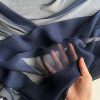 Une main présente le Georgette de soie au mètre de Longan Craft France - un drapé en crêpe de soie 100% mûrier, 8 mommes - révélant sa texture diaphane et fluide ainsi que sa translucidité, les doigts étant visibles à travers la douce soie bleu marine.
