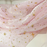 Gros plan sur le tissu organza étoilé au mètre de Longan Craft France, un tissu rose pâle brillant et translucide avec des motifs d'étoiles et de points dorés, drapé en plis souples - idéal pour les robes de mariée et les costumes.