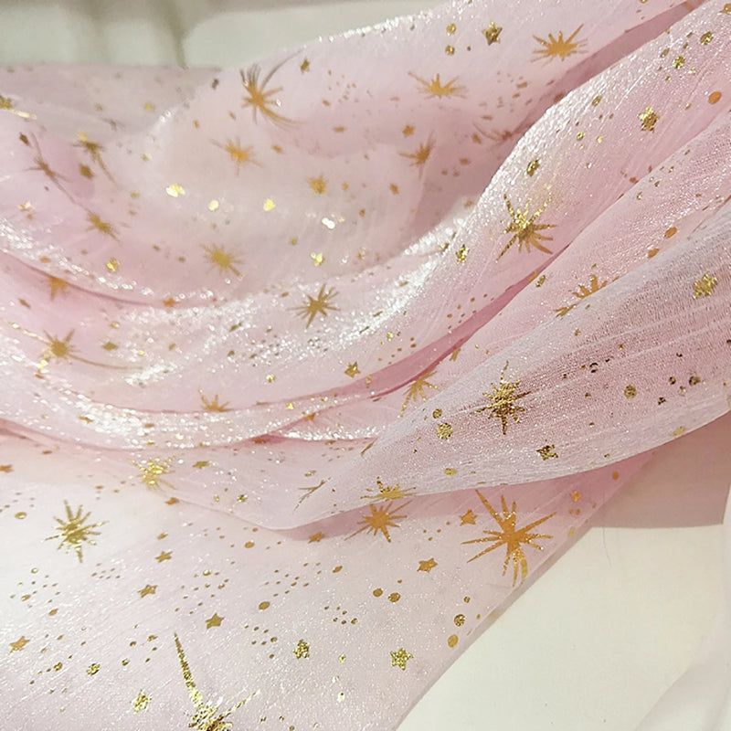 Gros plan sur le tissu organza étoilé au mètre de Longan Craft France, un tissu rose pâle brillant et translucide avec des motifs d'étoiles et de points dorés, drapé en plis souples - idéal pour les robes de mariée et les costumes.